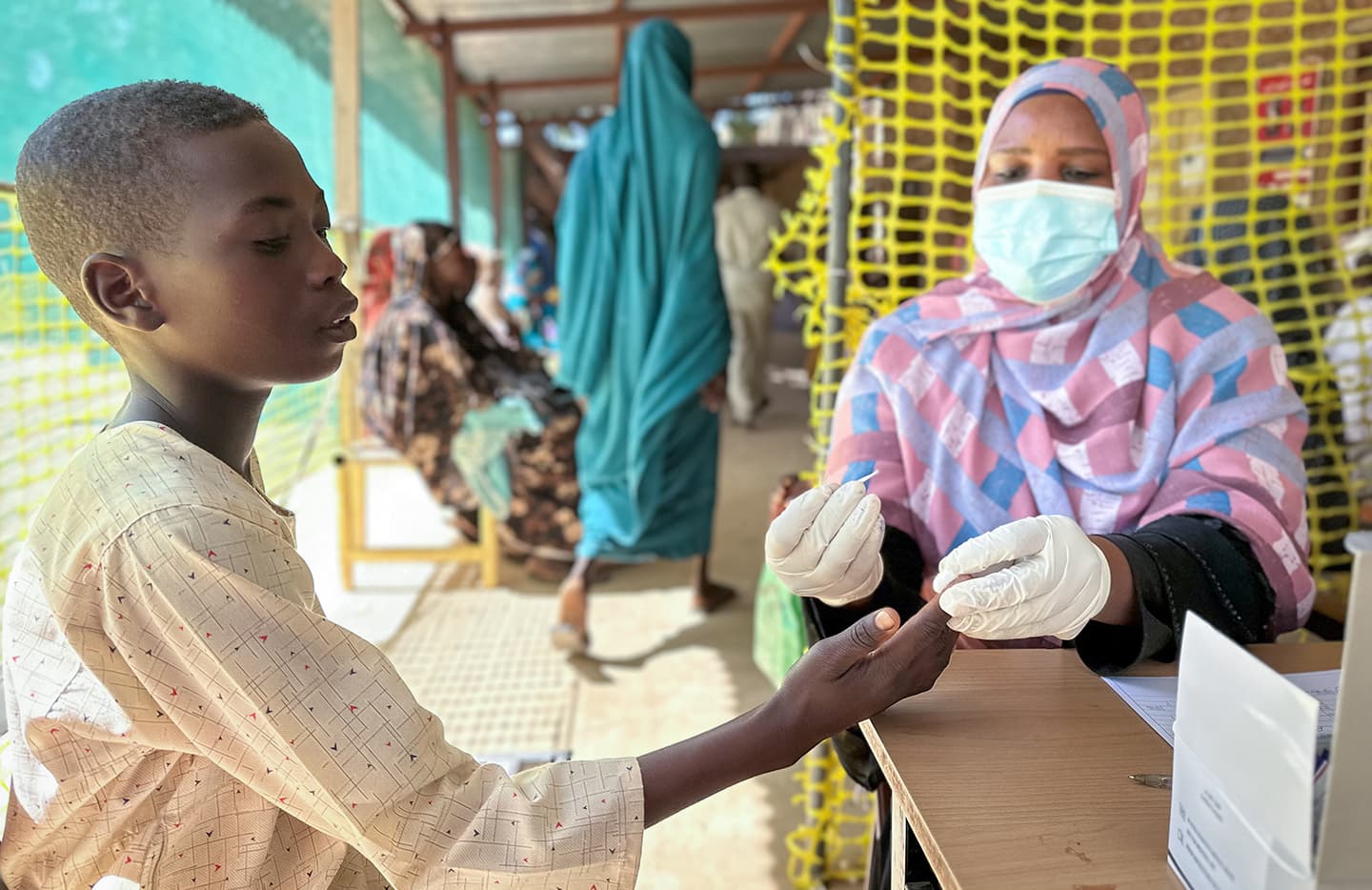 A healthcare worker checks a boy’s hand in an outdoor clinic. Other people wait in the background.